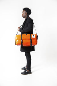 Sac Origami de la marque Dababo, sac pliable multi-fonctions, 3 positions en chutes de baches de camions recyclées porté de profil par un homme avec lanière en format fermé de couleur orange.