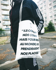 Totebag de la collaboration de Dababo avec le média lillois WWCF porté par un mannequin debout. Totebag en chutes de baches de camions recyclées avec la phrase "ils copient nos codes mais pour rien au monde, ils prendraient nos places". 