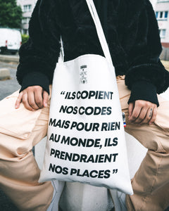 Totebag de la collaboration de Dababo avec le média lillois WWCF porté par un mannequin. Totebag en chutes de baches de camions recyclées avec la phrase "ils copient nos codes mais pour rien au monde, ils prendraient nos places". 